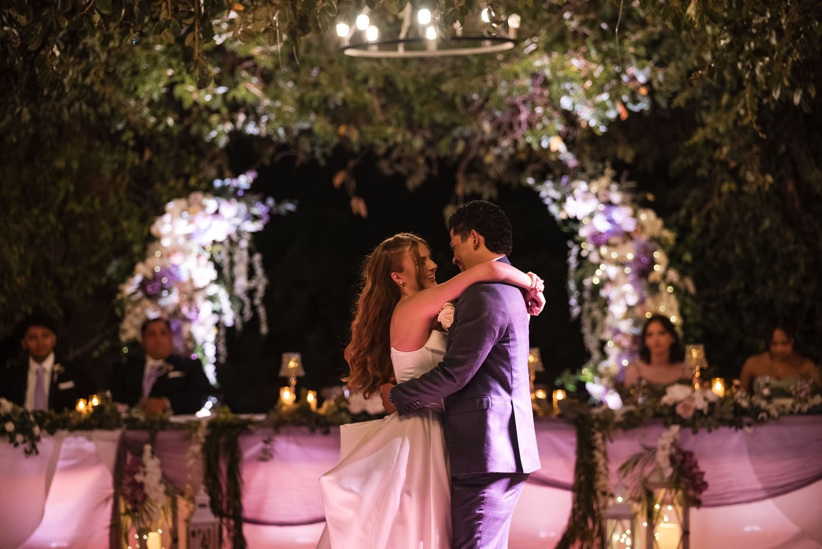 A first dance in front of the flower-lined head table under a chandelier