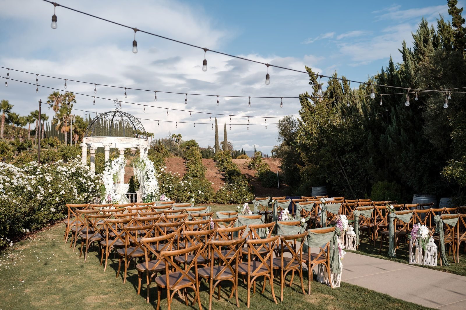 Rows of wooden chairs set for a garden ceremony at Europa Village in Temecula, string lights overhead and a white gazebo at the end of the aisle