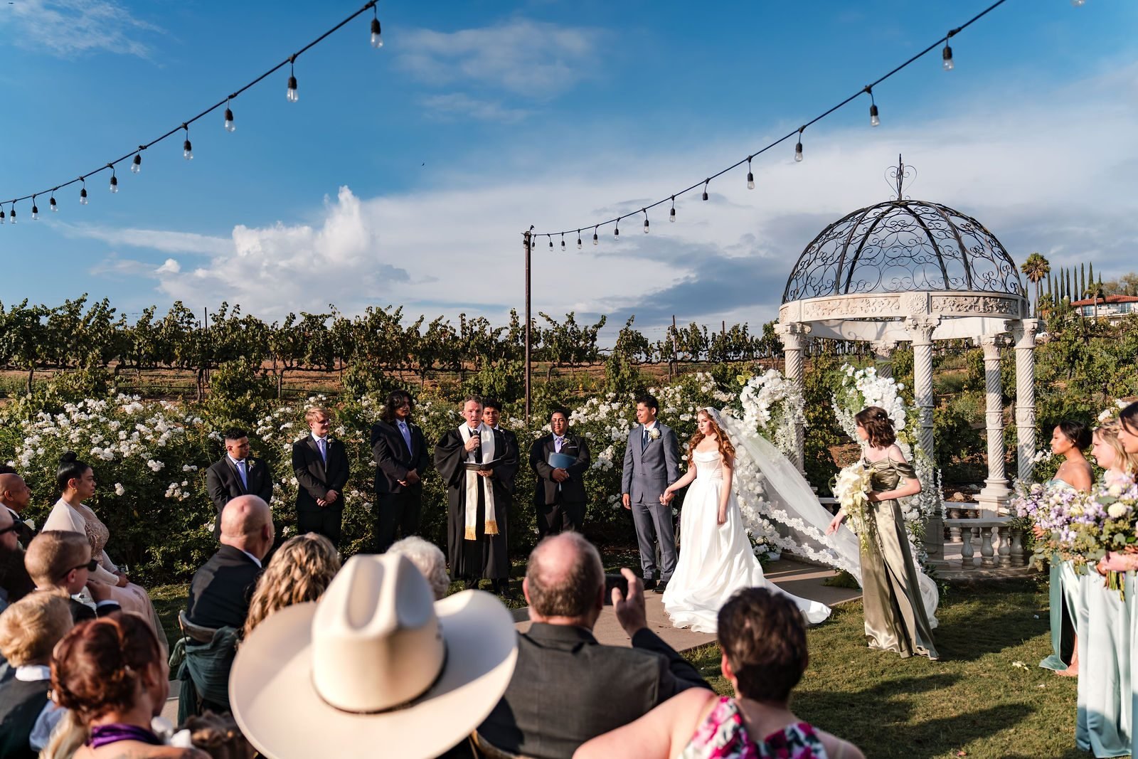 Guests watching a ceremony at a white gazebo at Europa Village, vines on the hill behind and string lights overhead
