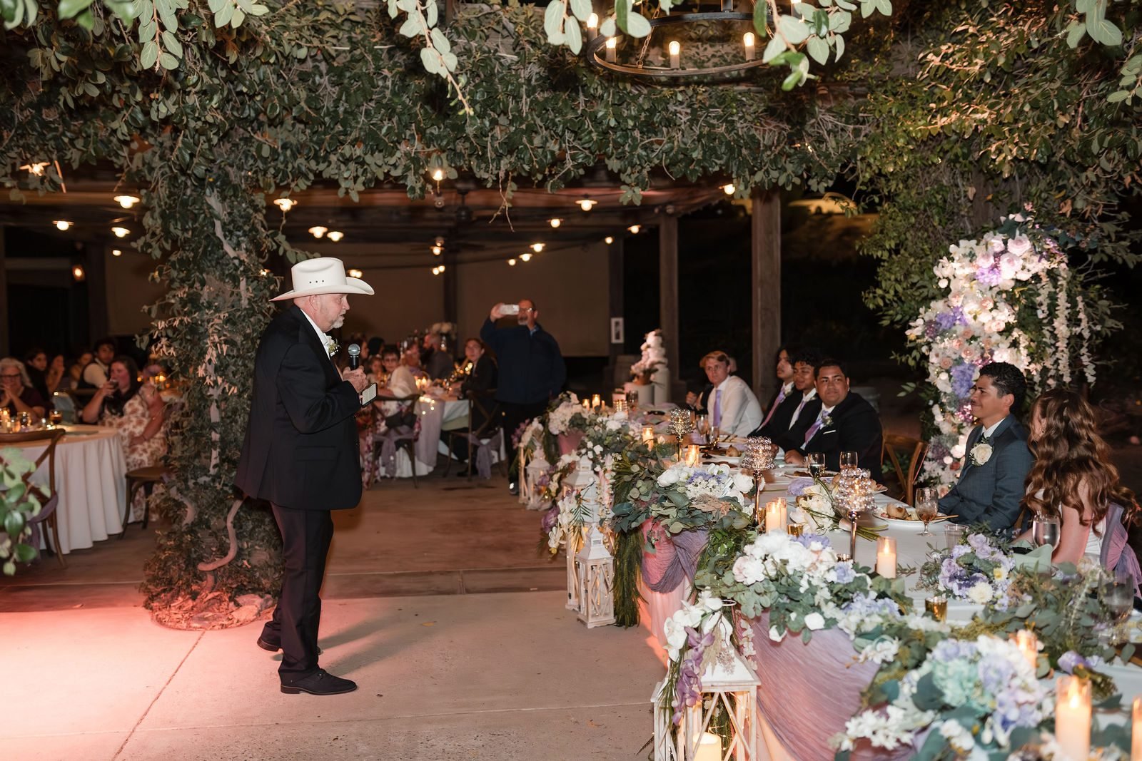 Reception tables under a canopy of trees and chandeliers, flowers along the head table