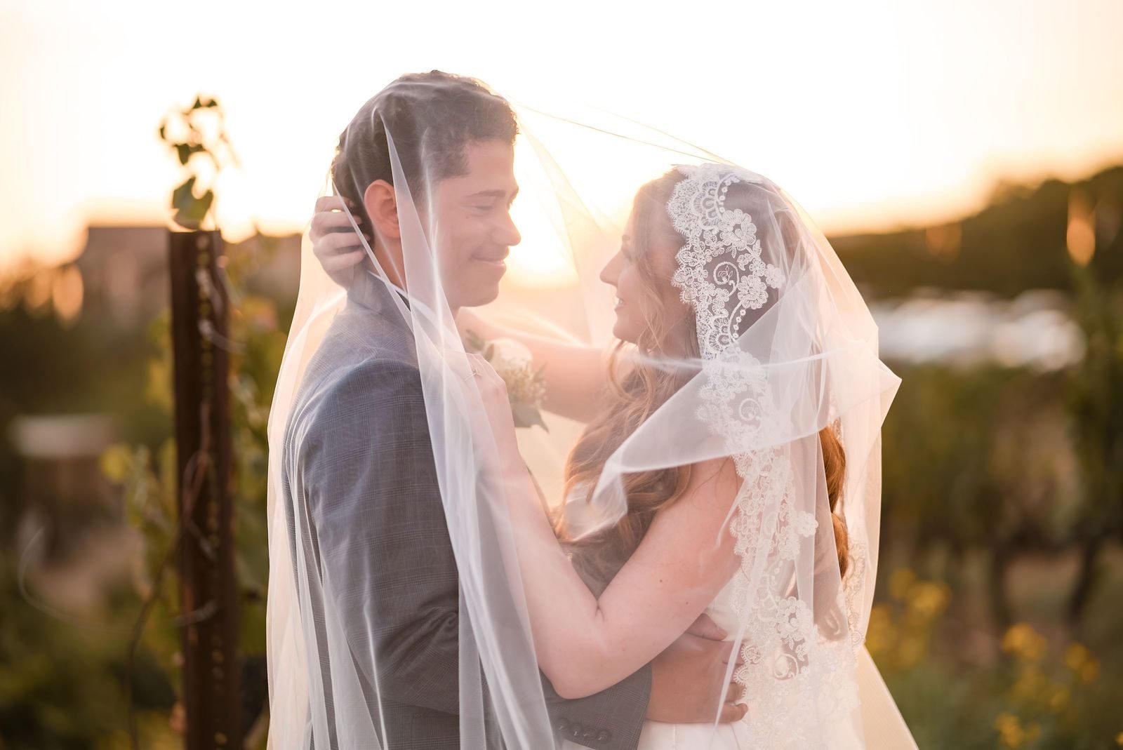 A couple close together under the bride's lace veil, vineyard posts lit by the late sun