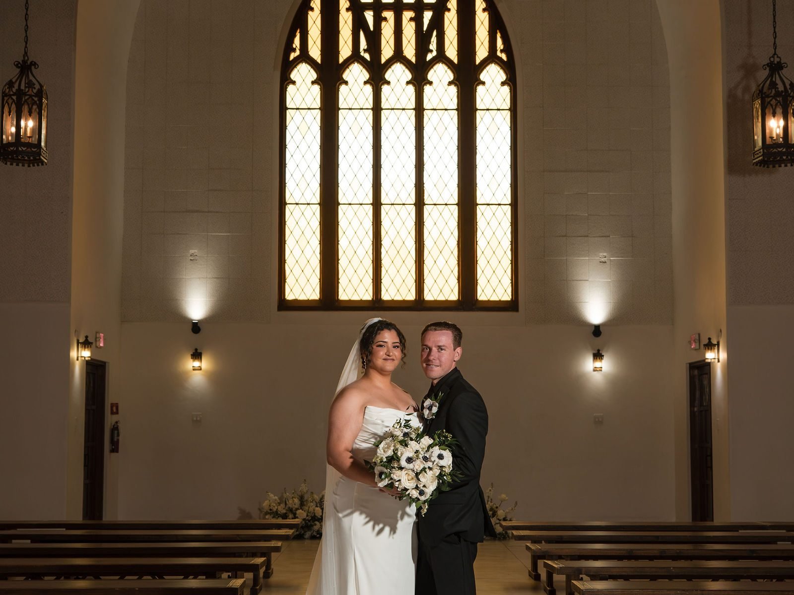 The bride and groom standing together beneath the tall arched window in the empty chapel
