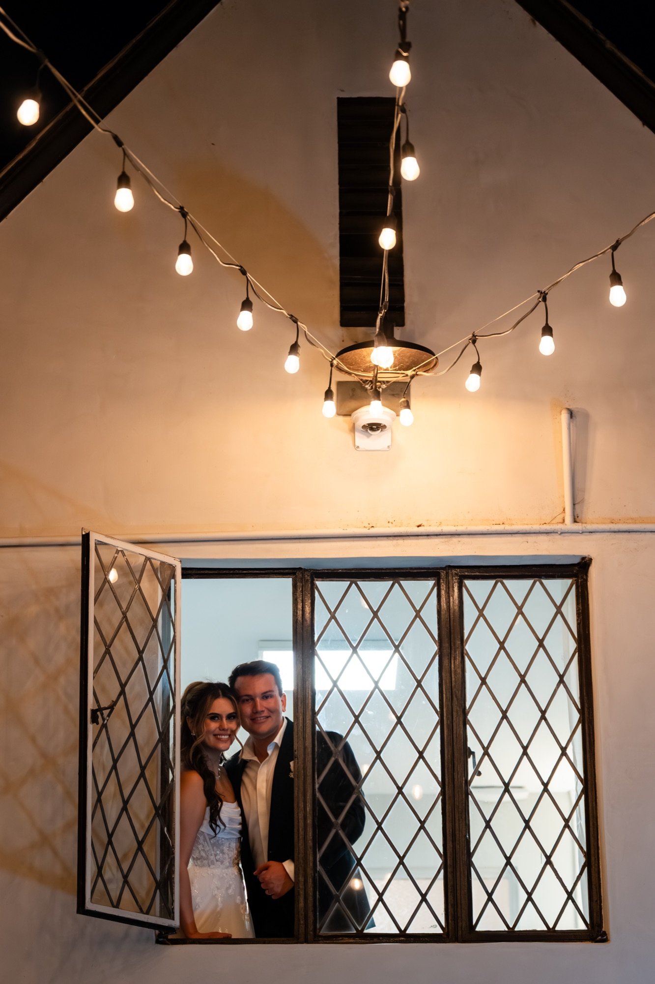 The couple leaning out of a diamond-paned window under string lights at night